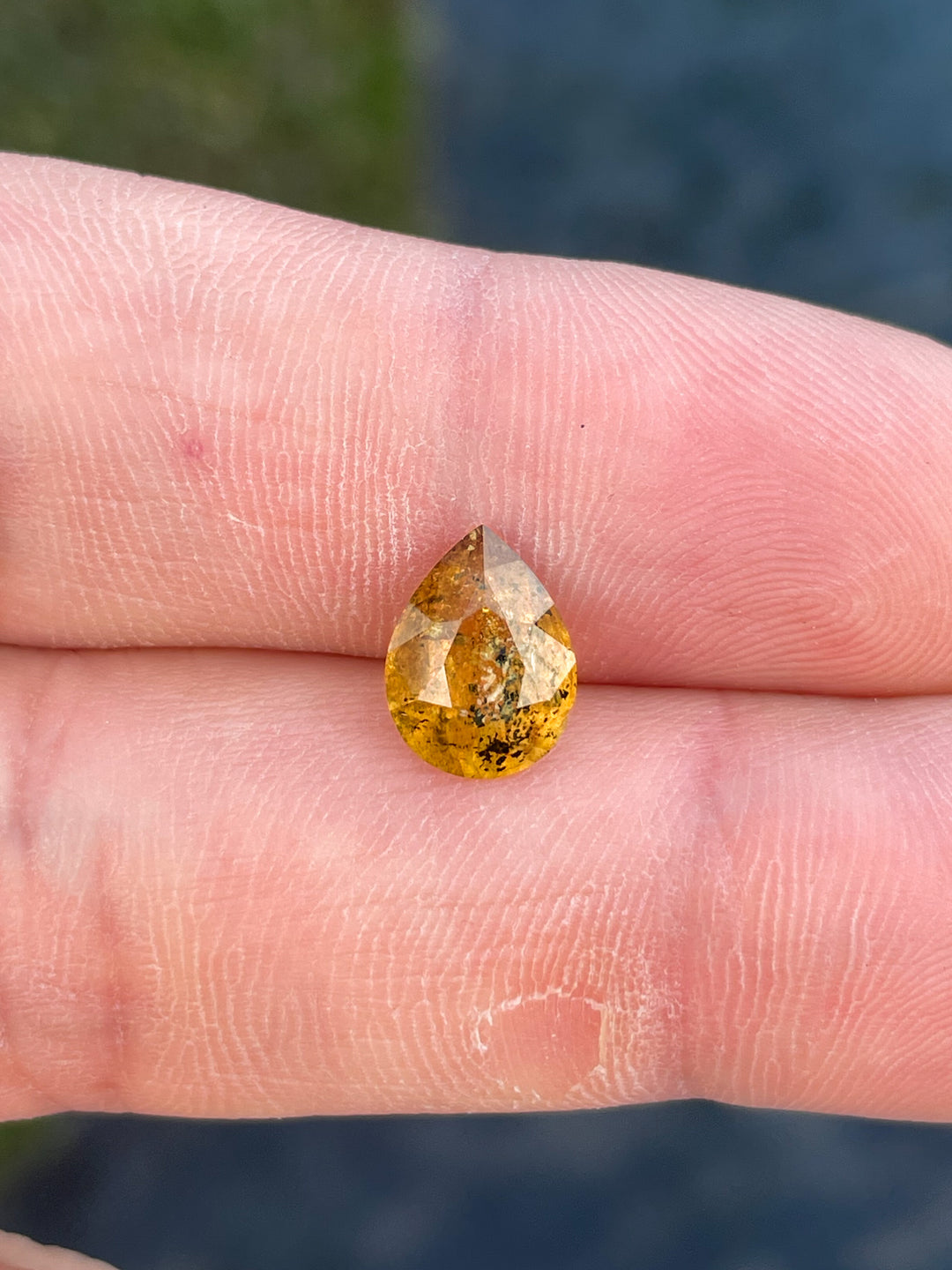 A richly hued sapphire with yellow and orange tones, resting on a finger, showcasing its pear-shaped modified cut.