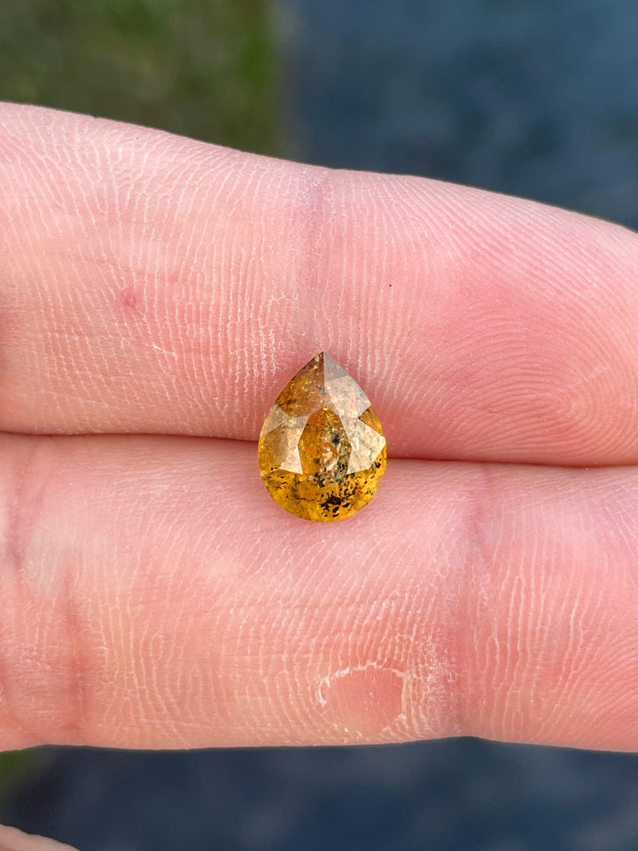 A richly hued sapphire with yellow and orange tones, resting on a finger, showcasing its pear-shaped modified cut.
