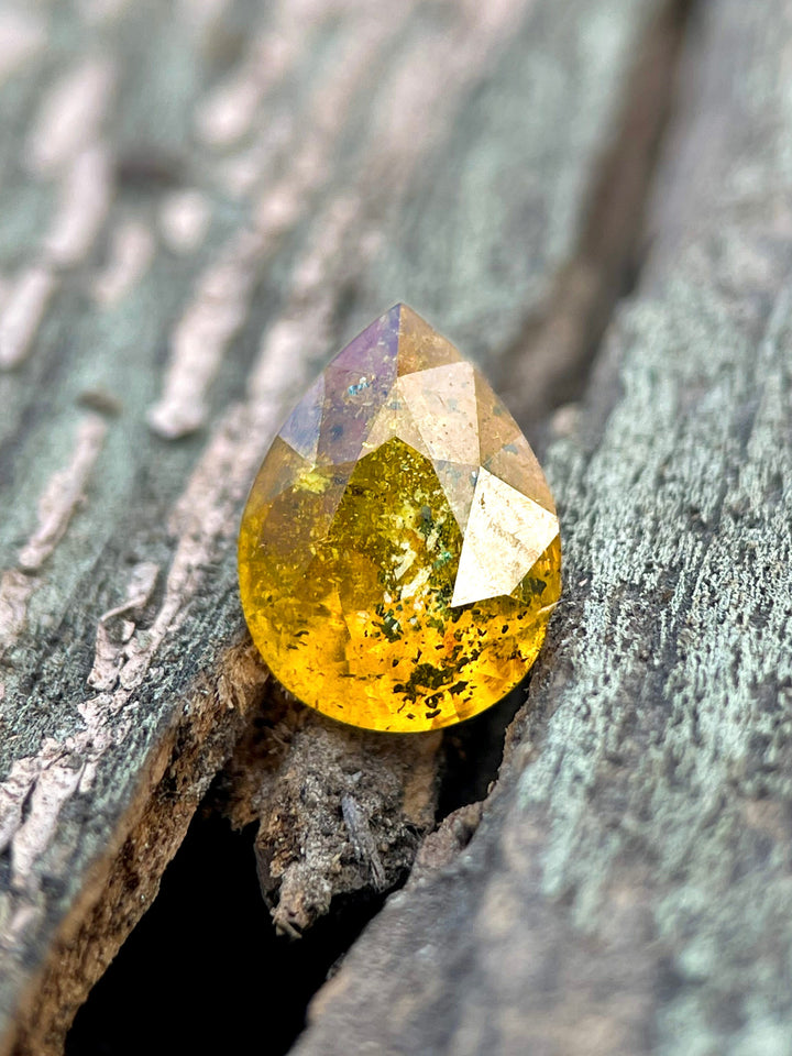 A 3.03-carat pear-shaped sapphire with a vibrant mix of yellow, orange, and black tones, placed on a rustic wooden surface.