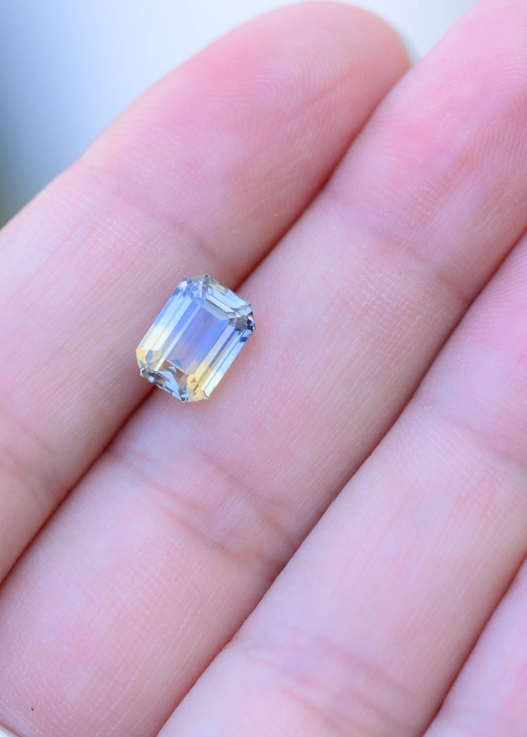 3.03 carat blue and yellow parti sapphire resting on fingertips for scale, showing its rectangular emerald-cut silhouette, transparent step facets and soft two-tone coloration.