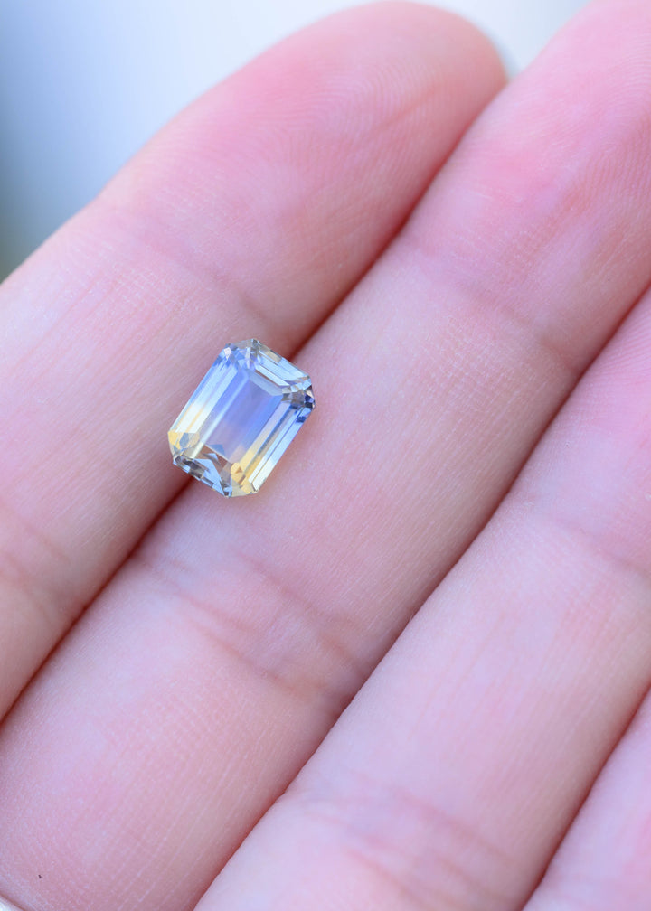 3.03 carat blue and yellow parti sapphire resting on fingertips for scale, showing its rectangular emerald-cut silhouette, transparent step facets and soft two-tone coloration.