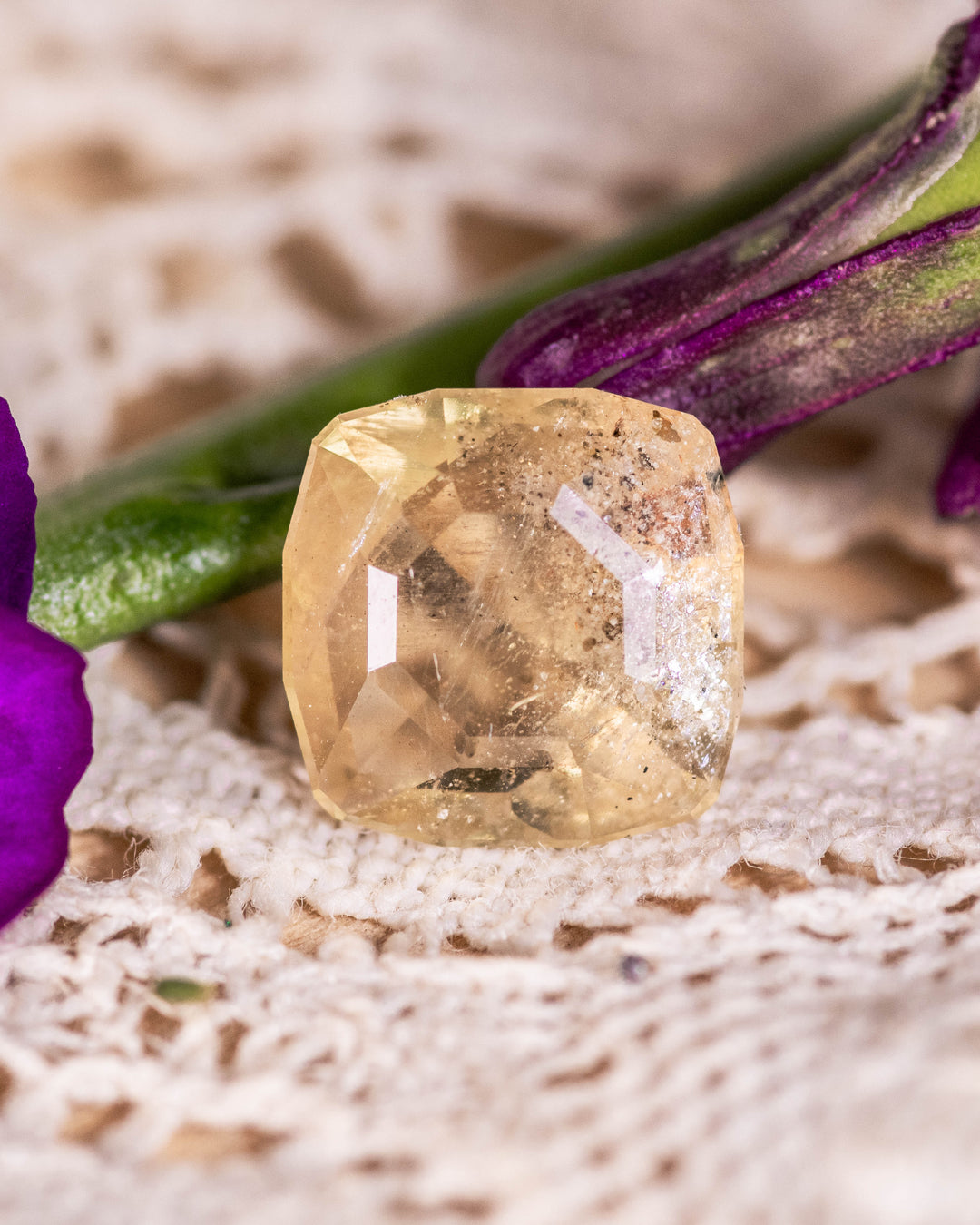 3.10 carat green yellow sapphire placed on vintage lace beside a purple flower, highlighting its natural earthy tones and custom cushion cut facets, untreated gemstone from Umba River, Tanzania.