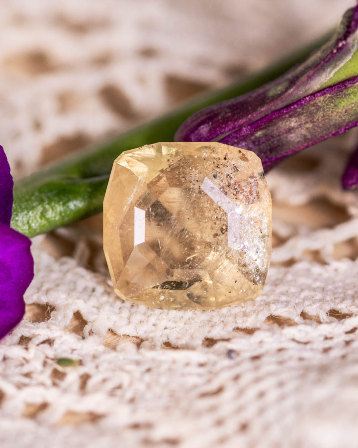 3.10 carat green yellow sapphire placed on vintage lace beside a purple flower, highlighting its natural earthy tones and custom cushion cut facets, untreated gemstone from Umba River, Tanzania.