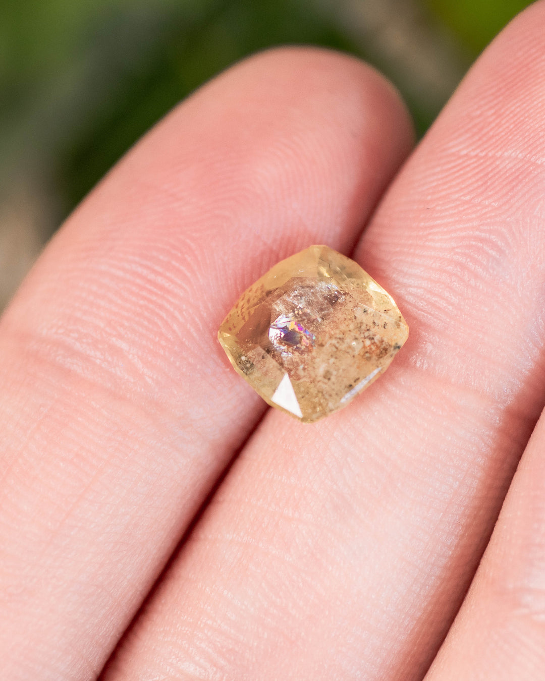 Macro image of 3.10 carat yellow green sapphire resting on fingertip, capturing brilliance, cushion cut symmetry, and organic inclusions of this untreated gemstone from Umba River, Tanzania.
