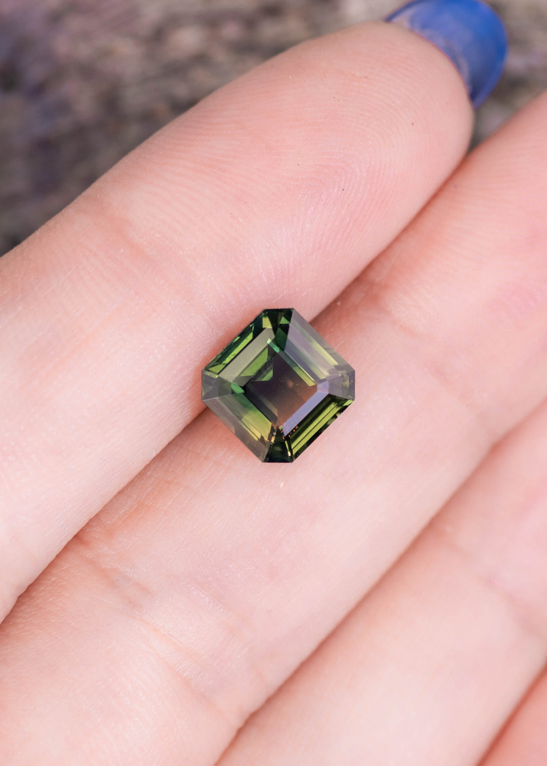 Loose 3.10 carat parti sapphire resting on a fingertip for scale, highlighting the square Asscher cut and the natural mix of green, yellow, and blue tones