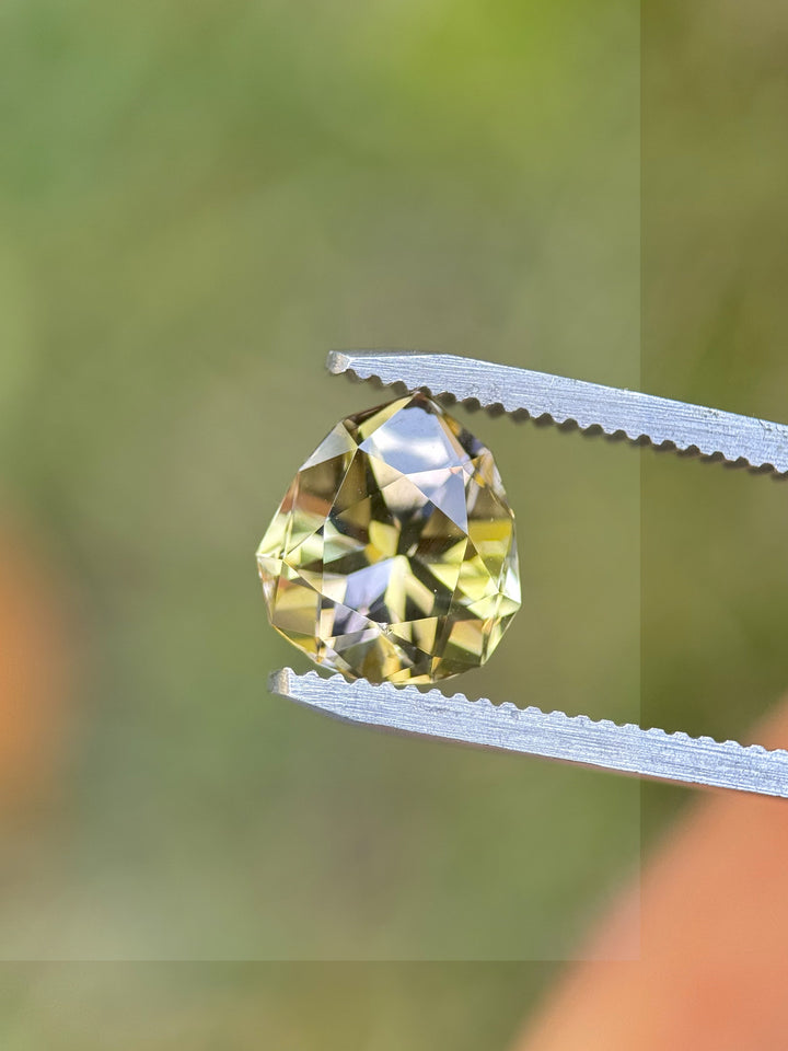 Golden yellow and lavender 3.25ct Tanzanite held in gem tweezers against a blurred green background. Custom pear mixed brilliant cut, no heat, from Tanzania’s Merelani Hills.