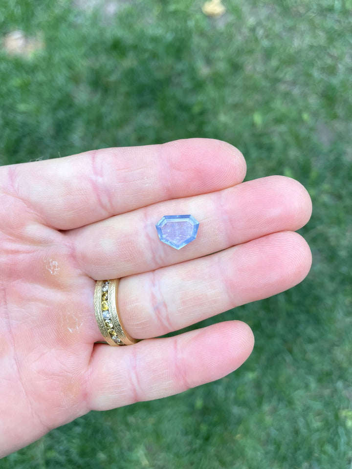 3.29 carat untreated sapphire resting on fingertips, displaying soft seafoam, teal, and blue hues in a custom step cut shield portrait shape.