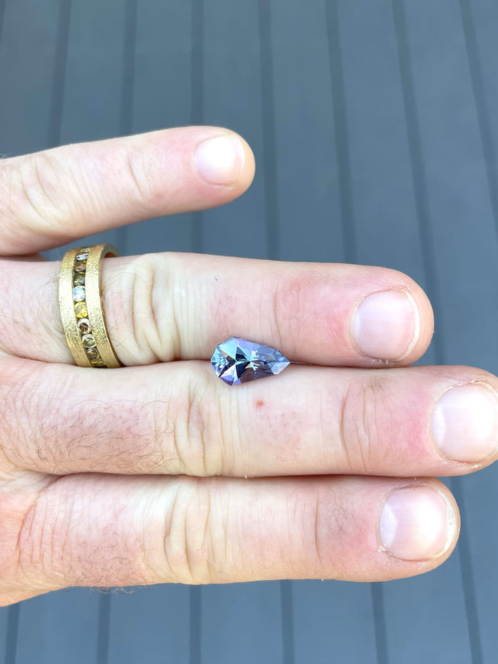 A 3.31-carat gray-blue-purple tanzanite gemstone displayed on a hand, showcasing its modified geometric pear cut and brilliant facets.