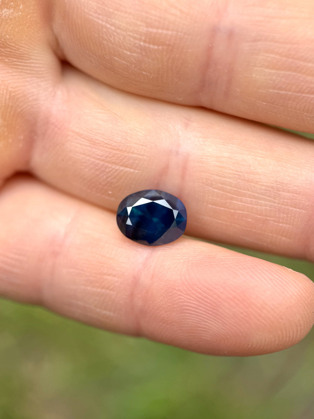 3.32-carat deep teal blue oval sapphire displayed on fingertips, showcasing its rich peacock color and brilliance.