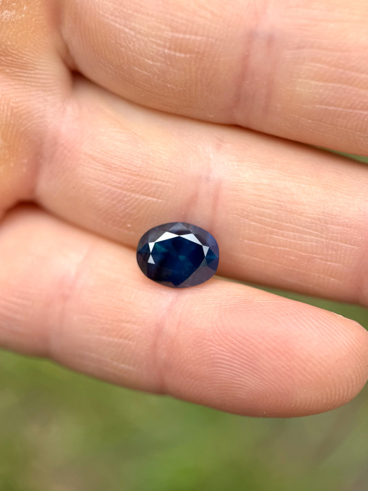 3.32-carat deep teal blue oval sapphire displayed on fingertips, showcasing its rich peacock color and brilliance.