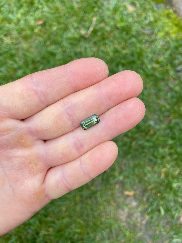 A 3.36-carat olive green opalescent sapphire from Gombe, Nigeria, displayed on a hand. The stone features a precision elongated emerald cut and subtle opalescent tones.

