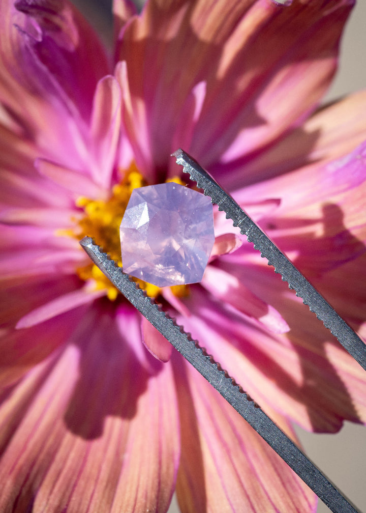 Opalescent lavender pink sapphire held in tweezers over a flower, showing the custom geometric radiant cut and soft Sri Lankan color.