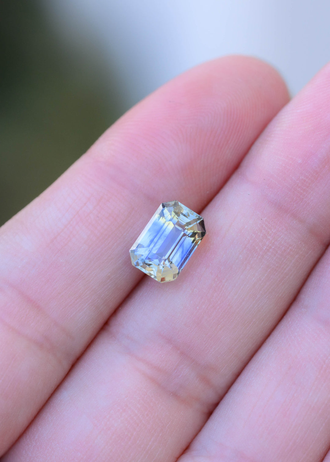 3.48 carat blue and yellow parti sapphire resting on fingertips for scale, showing its elongated rectangular proportions, transparent step facets and natural two-tone color zoning.