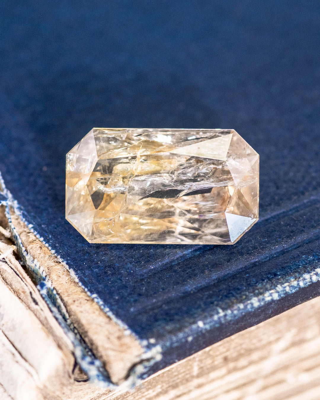 3.50 carat yellow orange sapphire resting on a deep blue book, showcasing radiant light reflections and intricate facets of its custom modified radiant cut, untreated gemstone from Umba River Valley, Tanzania.