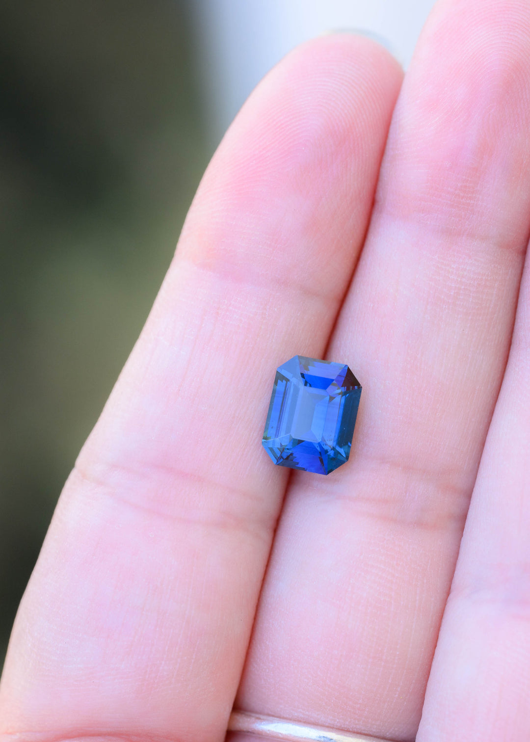 3.54 carat royal blue sapphire resting on fingertips for scale, showing its substantial 9.60 × 7.54 mm face-up size, emerald-cut geometry and subtle teal undertones.
