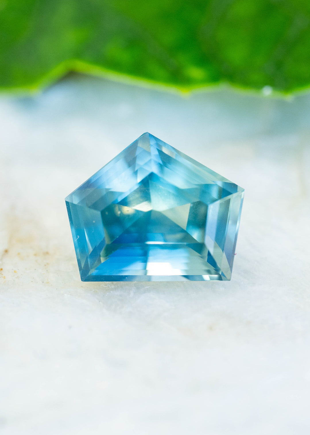 A 3.54-carat teal green blue sapphire from Diego, Madagascar, photographed on a pale natural surface. Its custom shield step cut reveals bright blue reflections, teal edges, and geometric facet patterns.