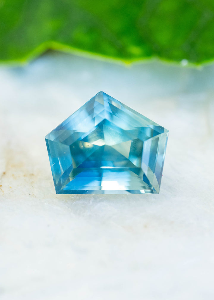 A 3.54-carat teal green blue sapphire from Diego, Madagascar, photographed on a pale natural surface. Its custom shield step cut reveals bright blue reflections, teal edges, and geometric facet patterns.