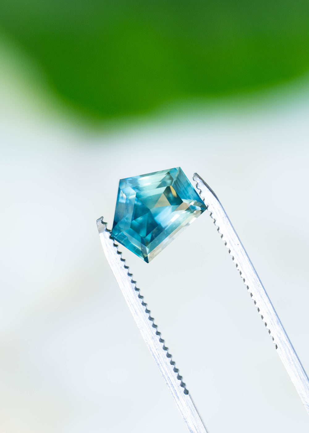 Close-up photograph of a 3.54ct heated Madagascar sapphire held in gem tweezers, highlighting its unusual shield outline, precision-oriented geometric faceting, and shifting teal blue color.