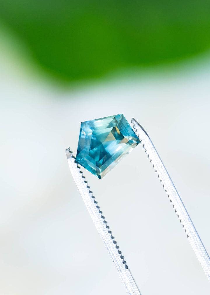 Close-up photograph of a 3.54ct heated Madagascar sapphire held in gem tweezers, highlighting its unusual shield outline, precision-oriented geometric faceting, and shifting teal blue color.