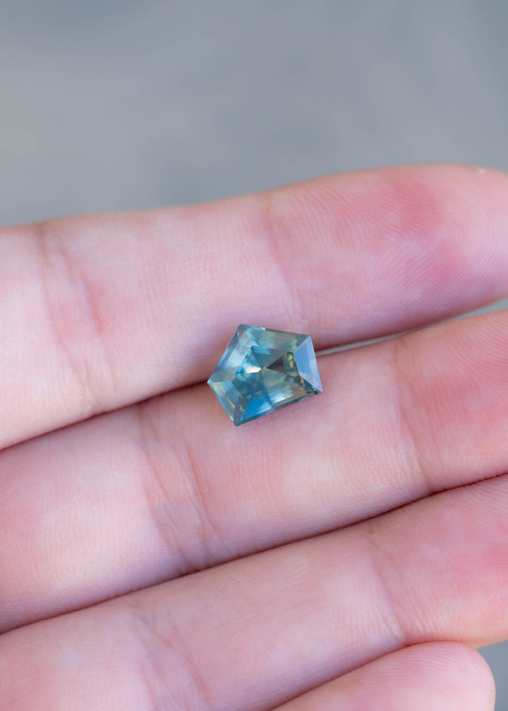 A 3.54ct custom shield step cut sapphire from Madagascar photographed on fingers to demonstrate its size, angular outline, and blue-green color in natural light.