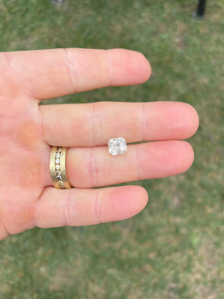 A pale yellow-gray-blue custom radiant-cut sapphire gemstone weighing 3.60 carats, held between fingers for scale.