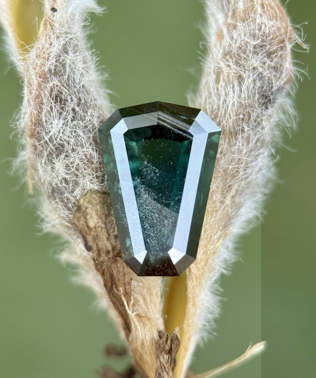 An untreated 3.65ct sapphire from Kenya with layered peacock, teal, and blue color, resting on a textured stone surface with earthy brown and tan hues beneath it.