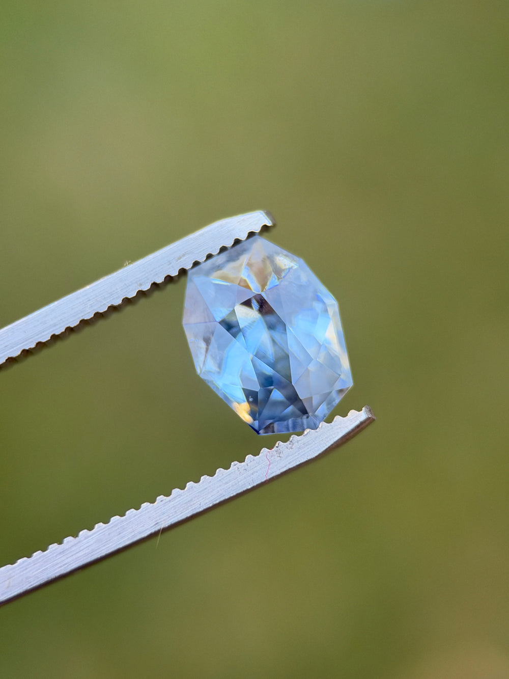 Natural Sri Lankan sapphire held in tweezers showing pale sky blue, deeper denim blue zoning and geometric shield facets