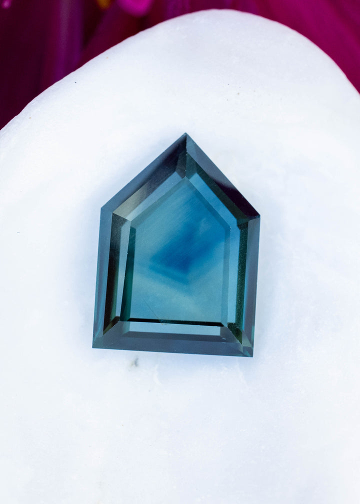 Five-sided shield-cut sapphire resting on a white flower petal against a magenta floral background. The stone has a misty blue center framed by dark teal-green facets.