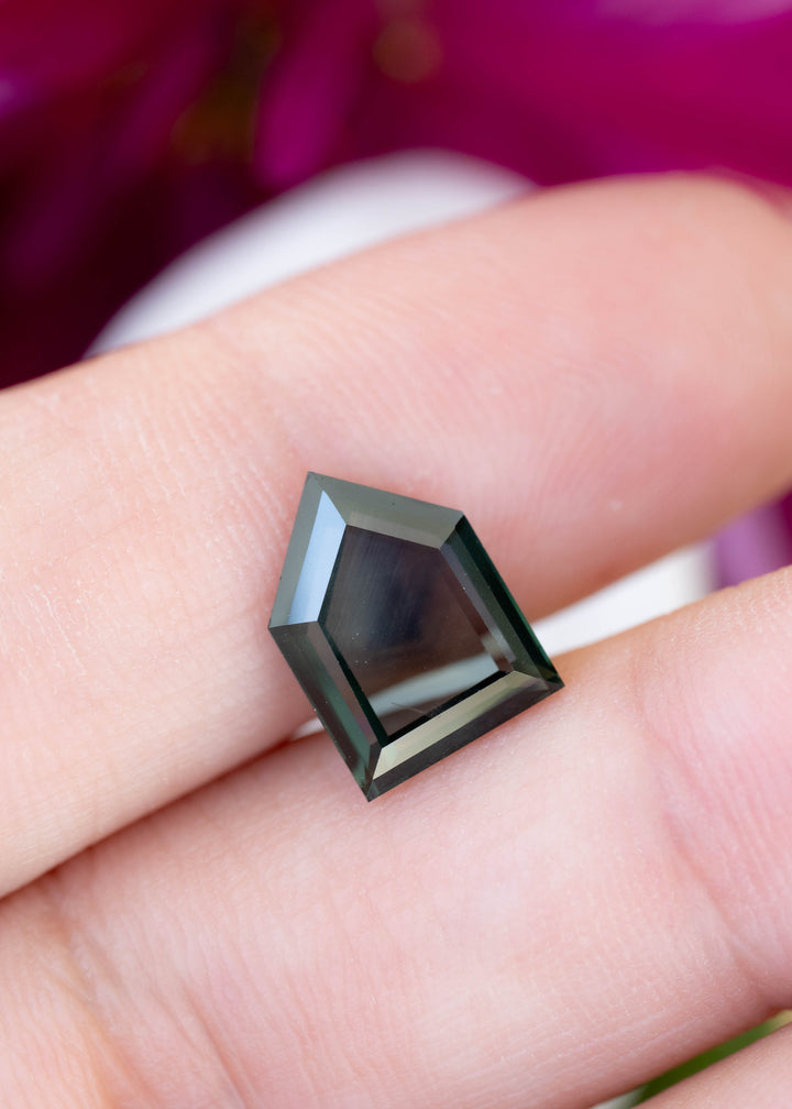 Dark teal-green five-sided sapphire resting across two fingertips. Its broad central facet appears smoky and reflective, with lighter blue-green reflections along the stepped edges.