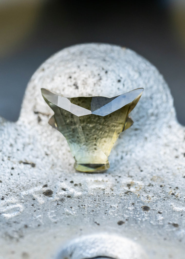A faceted gemstone with a triangular shape, showing a color gradient from olive green to pale yellow, resting on a textured gray stone surface.