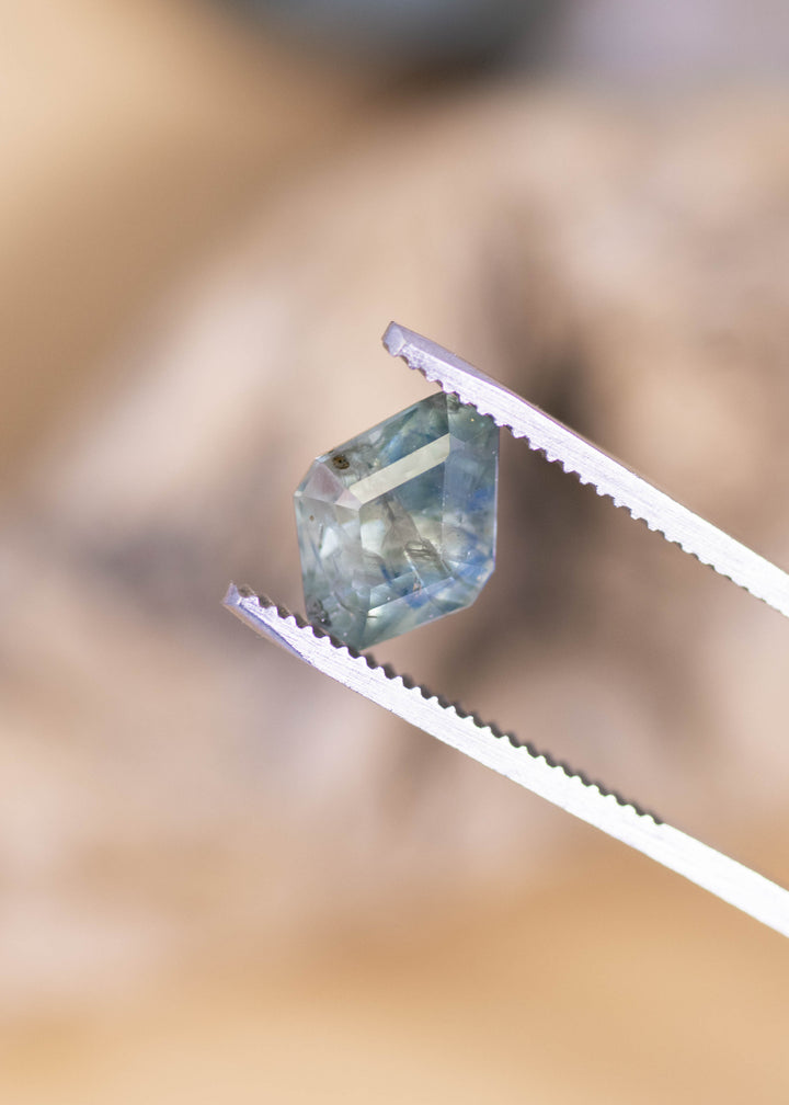 Geometric kite-shaped Montana sapphire held in silver tweezers, displaying layered teal and navy blue color bands with visible internal silk and natural inclusions under direct light.