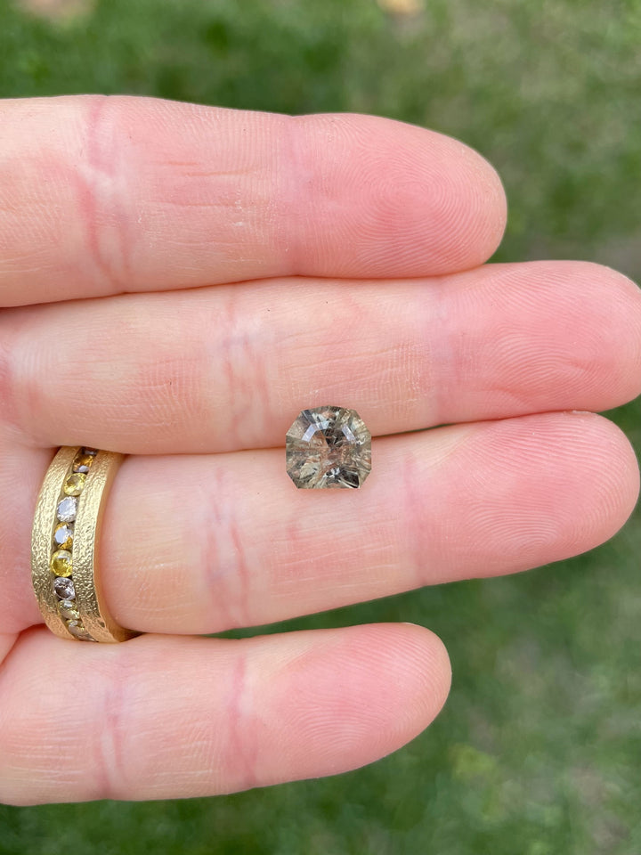 Hand holding a 3.82 carat color-changing sapphire from Umba River, Tanzania, showing earthy grey-green hues.