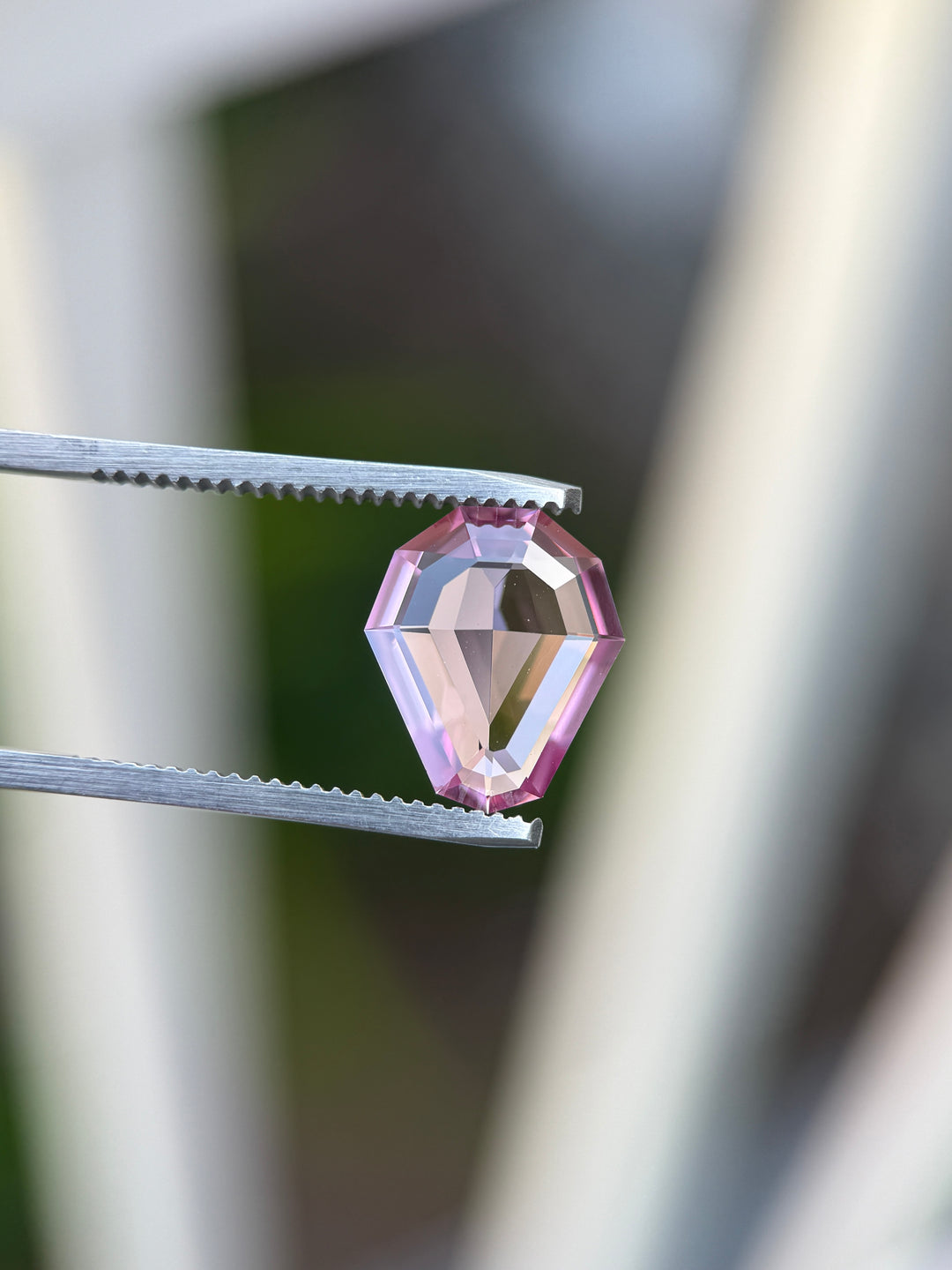 A faceted pink gemstone held between metal tweezers against a softly blurred outdoor background.
