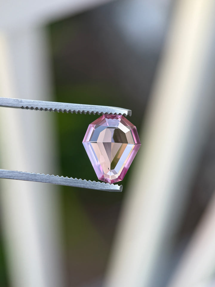 A faceted pink gemstone held between metal tweezers against a softly blurred outdoor background.