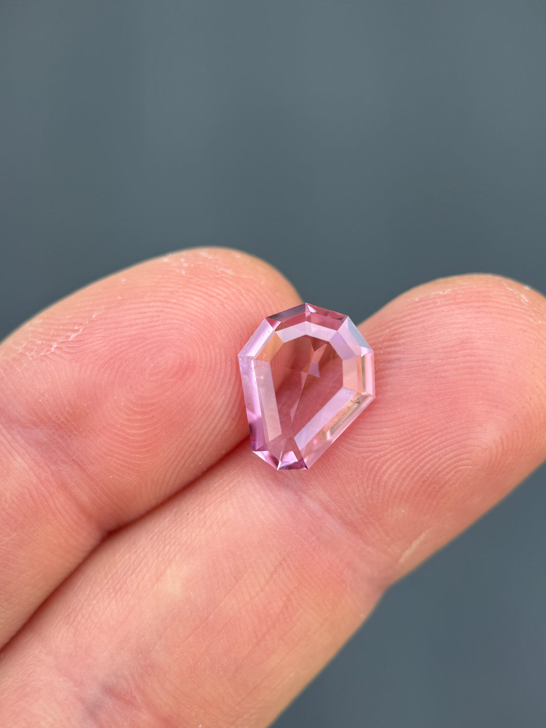 A faceted light pink gemstone held between two human fingers against a dark gray background.