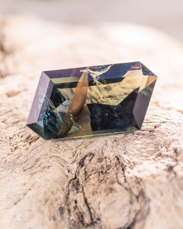 Close-up of 3.93 carat teal blue green sapphire resting on natural driftwood, reflecting metallic peacock hues and intricate facet design of its custom corner rhombus mixed cut, untreated sapphire from Garba Tula, Kenya.
