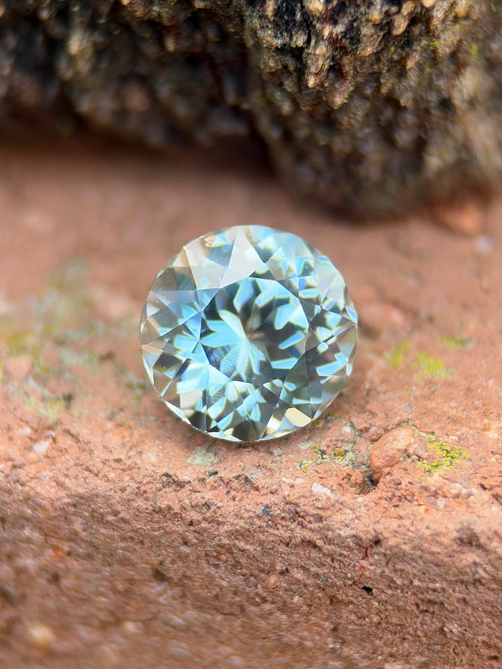 A round brilliant-cut light blue sapphire resting on a textured reddish-brown surface with a blurred natural rock background.