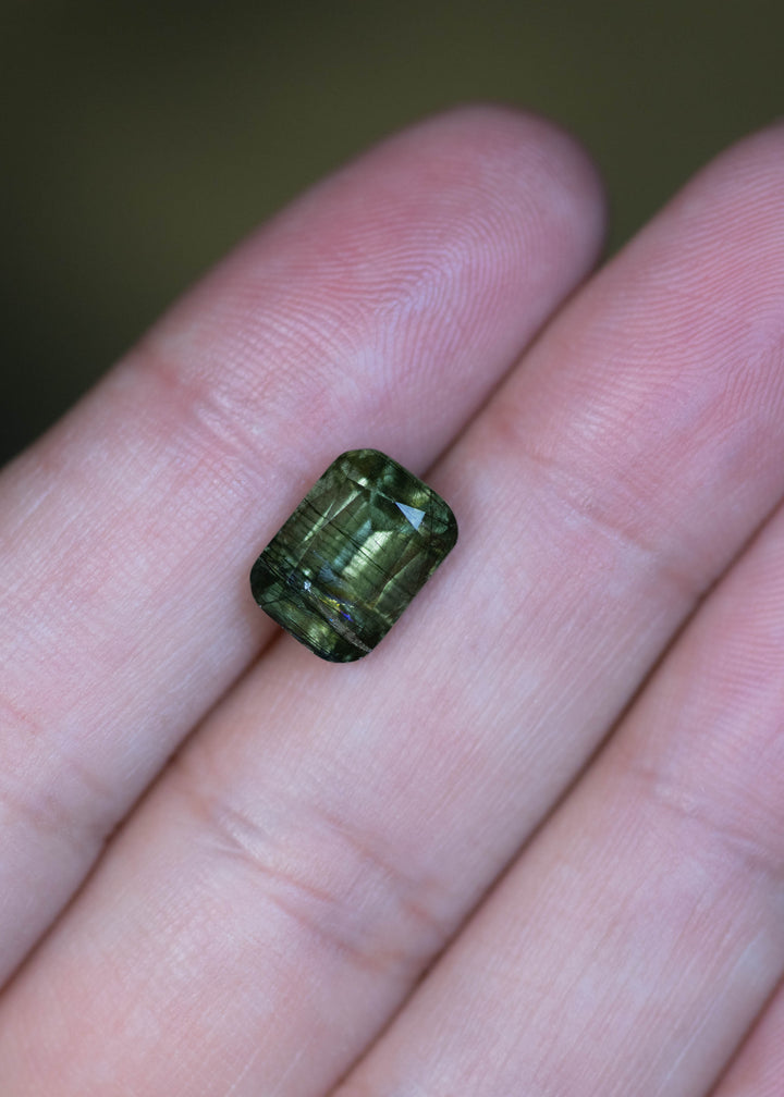 Natural untreated 4.35 carat forest green peridot from Skardu, Pakistan shown on fingertips for scale, displaying its cushion mix cut and earthy green tones.