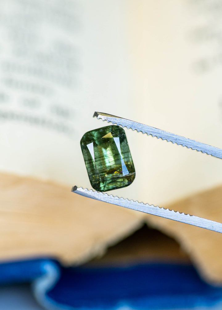 Close-up of a natural forest green Pakistan peridot held in tweezers, showcasing its custom cushion mix cut and intricate natural crystal inclusions.