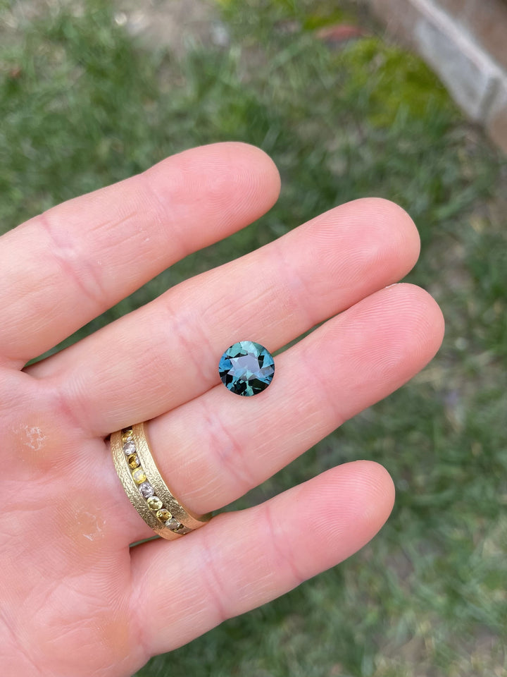 Hand holding a 4.00-carat round antique brilliant cut sapphire with peacock, blue, and green hues from Diego, Madagascar