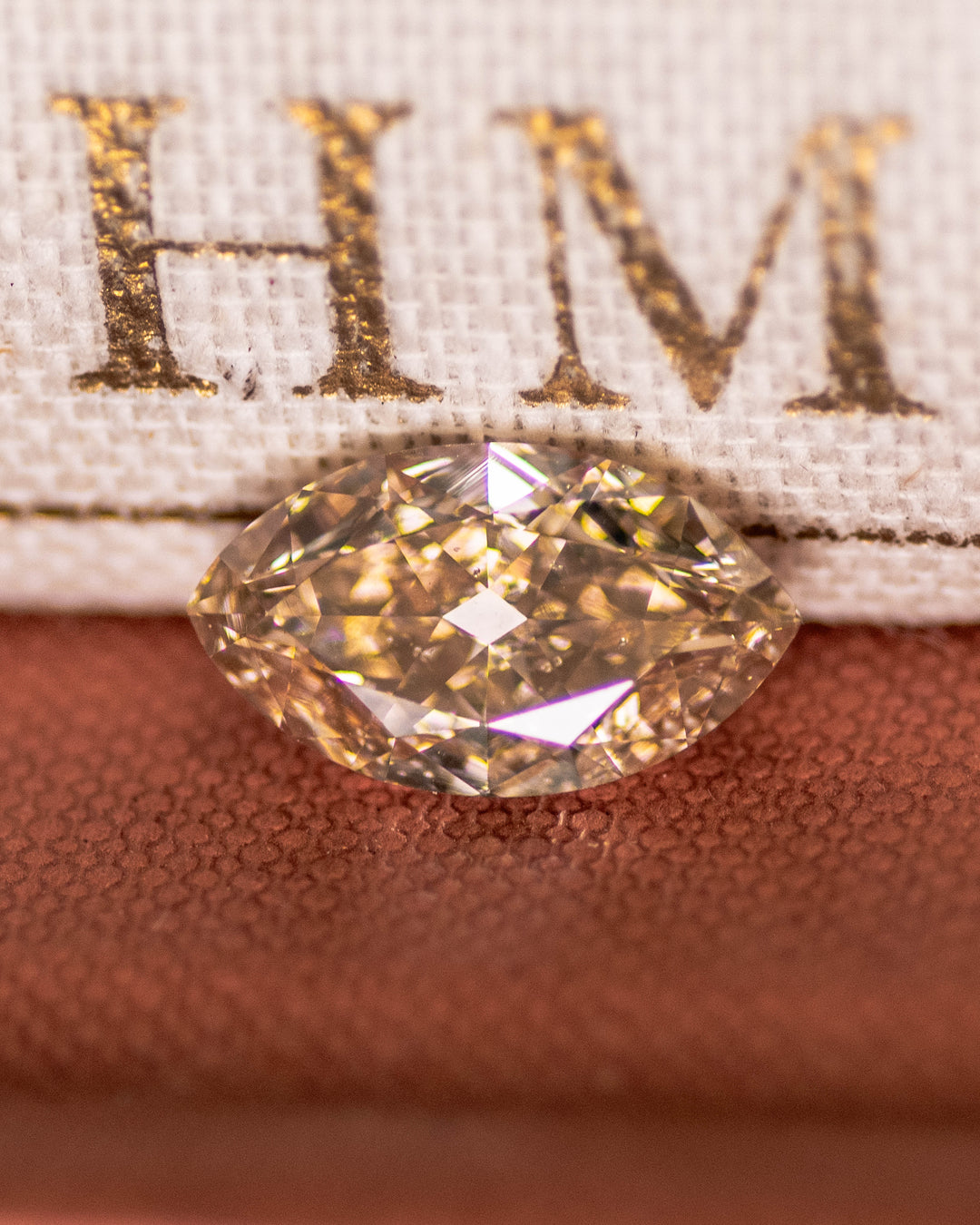Macro shot of a 4.01 carat rosy champagne fancy yellow brown diamond resting on a textured book cover. The antique-style moval modified brilliant cut from South Africa displays warm champagne hues and strong blue fluorescence.