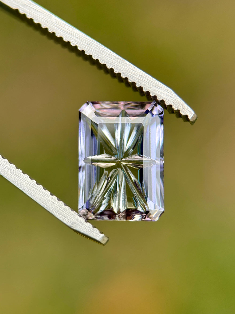 4.01CT Tanzanite held in gem tweezers under natural light, revealing blue, pink, and purple multi-color shifts and the custom concave fantasy emerald cut. Untreated Merelani Hills Tanzanite with sharp internal reflections.