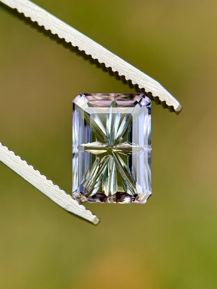 4.01CT Tanzanite held in gem tweezers under natural light, revealing blue, pink, and purple multi-color shifts and the custom concave fantasy emerald cut. Untreated Merelani Hills Tanzanite with sharp internal reflections.