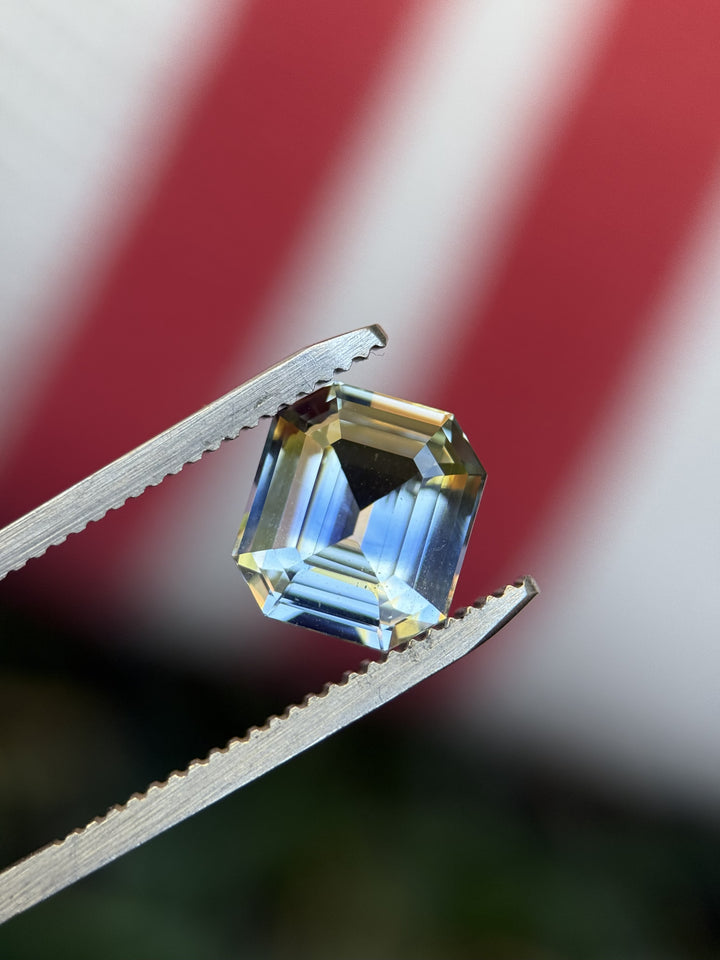 Precision emerald cut 4.04CT parti sapphire held in gem tweezers against a striped red and white background, displaying its light yellow and blue color zoning and untreated clarity.