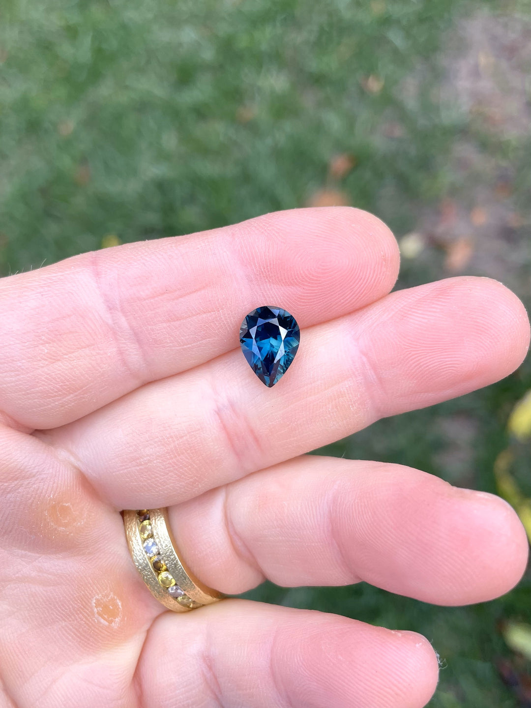 4.20 carat untreated sapphire displayed on fingertips with a grassy background, showing vibrant green, blue, and teal hues in a pear cut.