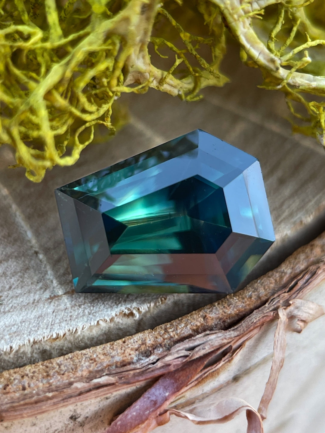 A stunning 4.65-carat sapphire with forest green, teal, and yellow hues in a step cut coffin shape, resting on a textured natural surface.