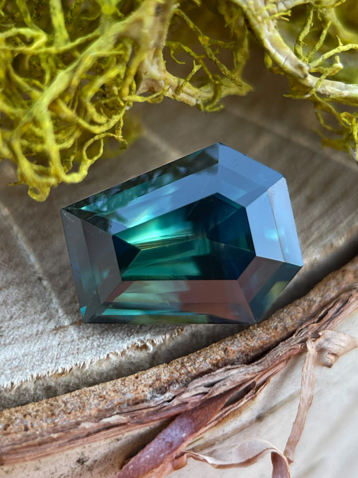 A stunning 4.65-carat sapphire with forest green, teal, and yellow hues in a step cut coffin shape, resting on a textured natural surface.