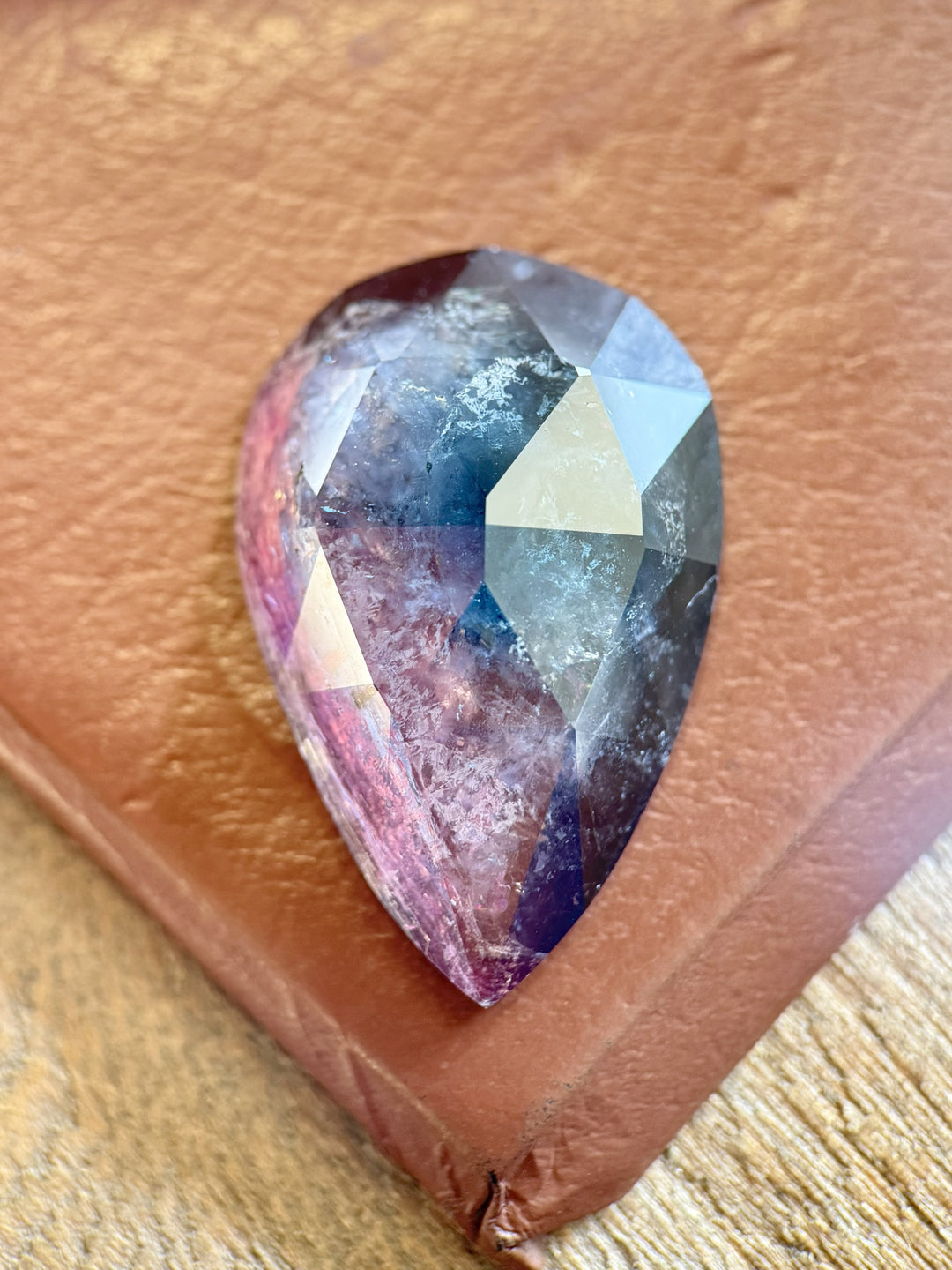 46.80 carat bi-color tourmaline with purple, pink, and blue hues resting on a textured brown surface, showing its custom pear rose cut facets and internal crystal formations, untreated and sourced from Paprok Mine, Afghanistan.