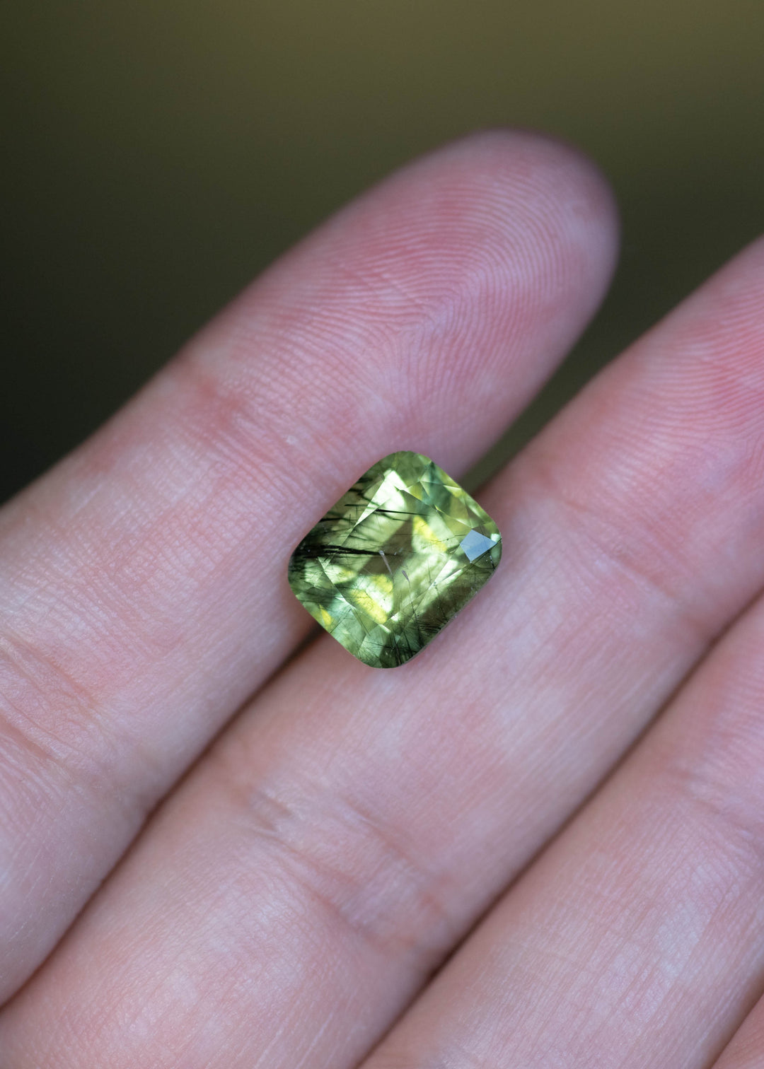 Natural untreated 5.49 carat Pakistan peridot shown on fingertips for scale, displaying its cushion mix cut and soft sage green coloration.