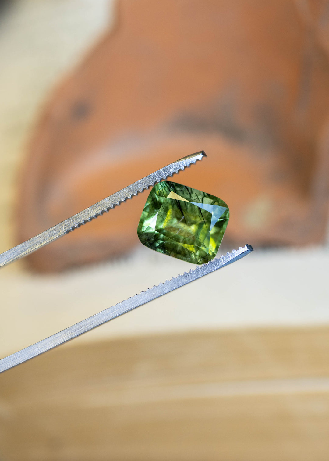 Close-up of a natural Pakistan peridot held in tweezers, showcasing its cushion mix cut, soft sage green color, and intricate crystal inclusions.